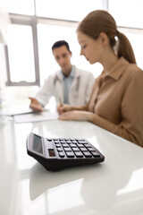 Fototapeta premium Closeup calculator lies on desk as patient consults with doctor in background, discussing charges and reviewing treatment-related documents, signing contract, get insurance coverage of private clinic
