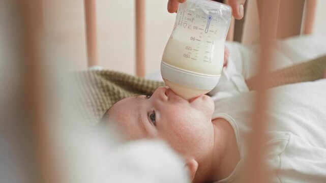 Baby boy drinking from bottle with mouth latched in crib inside building nursery, rails visible; tender calm serenity.