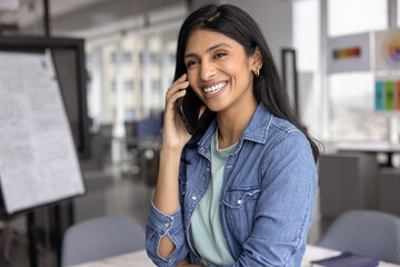 Fototapeta premium Attractive Indian businesswoman smiling, enjoy pleasant personal or business-related conversation on smartphone while stands alone in modern workplace. Remote communication, workflow using technology