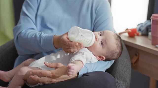 Newborn boy fed from a bottle while caregiver holds him and offers milk at a chair in a building; comfort bonding.