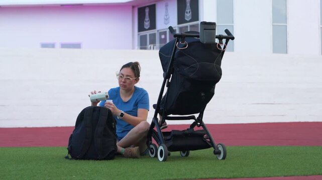 A young Latina mother carefully prepares baby formula from a thermos next to a stroller on artificial grass by the modern Mega Escultura building in Chetumal. The scene captures the daily life of a ne