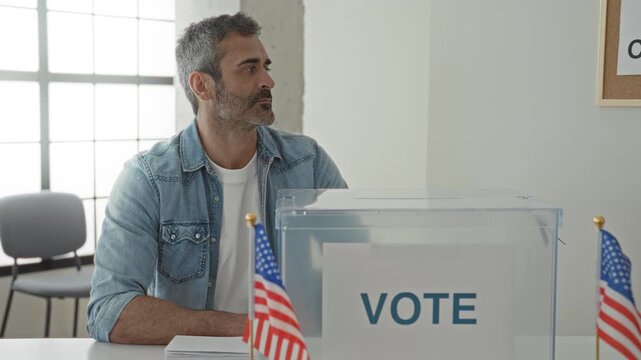 Man sits behind a clear ballot box at a polling table with small us flags and a stack of ballots inside a government building; seriousness.