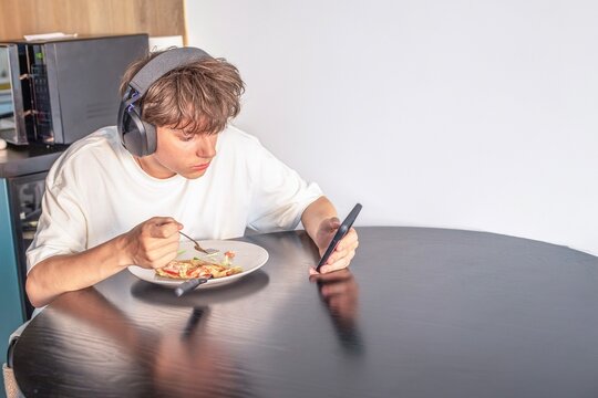 Teenager boy in headphones looking at smartphone while eating lunch at home