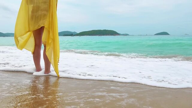 Female legs in yellow pareo standing at shoreline. Foamy ocean waves wash over bare foot. 