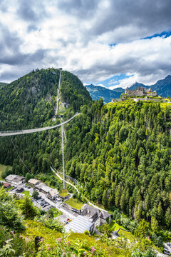 The view shows a suspension bridge connecting to the Gothic Ehrenberg Castle and the Schlosskopf fortress in Austria. Mountains surround the area, and trees cover the landscape.