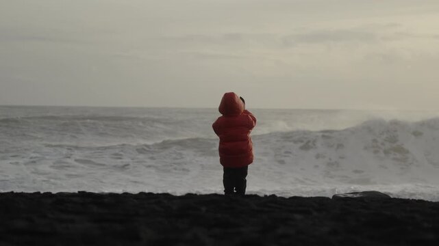Slow motion shot of a child standing on the black sand beach of Reynisfjara with powerful waves in the background during a beautiful sunset in Iceland