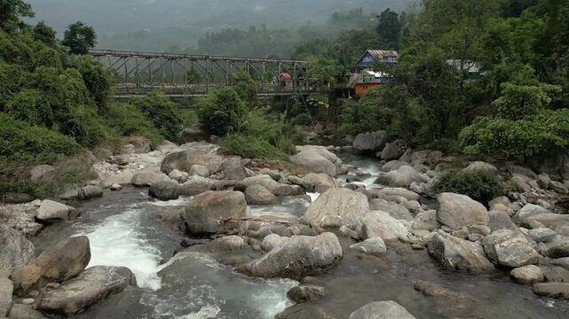 A cinematic drone shot of a rocky mountain river flowing under a vintage iron bridge in Sittong, North Bengal, India. nature scene for travel.