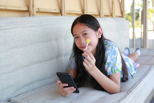 Portrait of young Asian girl lying down on cozy outdoor sofa, smiling happily while holding a smartphone and a yellow lollipop in her hands.