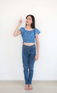 Portrait lovely young Asian girl in blue denim crop top and matching blue jeans, smiling while pointing her index finger up and looking upward against white wall background.