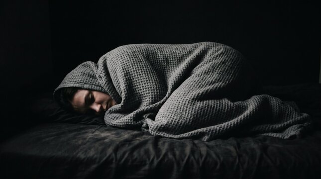 Person curled up under a blanket in a dark room, feeling sad.