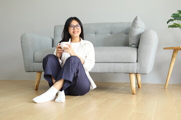 Obraz premium A happy young woman holding a cup of coffee while relaxing sitting on the floor leaning on the sofa at home, smiling while looking to the side.