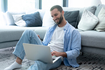 Smiling man sitting on living room floor using laptop, relaxed casual remote work at home,...