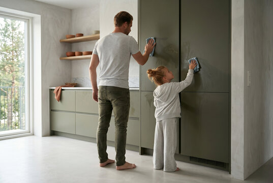Three-quarters back view of a father and daughter working in unison, tracking their cloths across a tall, integrated dark olive-green kitchen unit in soft directional daylight.

