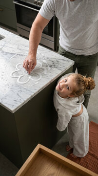 High-angle top-down shot of a father tracing a soap flower on veined marble, while his daughter looks up directly into the camera with delighted eyes by the olive island base.
