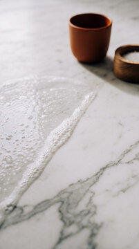 A macro view of a white marble tabletop split diagonally: one half wet with glistening soap suds and tiny bubbles, the other half perfectly wiped dry, showing smooth stone structure.
