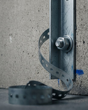 Vertical portrait of a galvanized steel angle bracket secured by a wedge anchor against concrete. The elegant curve of the perforated tape creates dynamic depth and industrial sophistication.