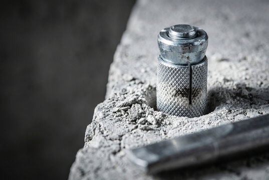 Extreme close-up of a knurled drop-in steel anchor inserted into a concrete hole. Powdery dust and hammer impact marks emphasize the raw, tangible nature of industrial installation.