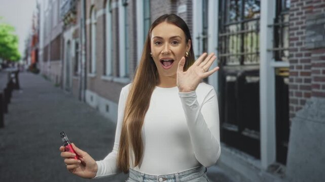 Woman waving hand while holding a red vaporizer on a city street with brick facades and sidewalk, mouth open in a wave; friendly greeting.