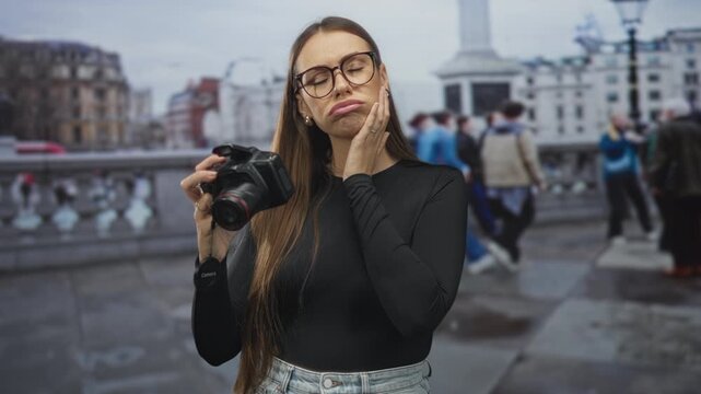 Woman holding dslr camera, touching cheek with palm, pensive pout and inspecting lens in crowded city street near monument; concern toothache.