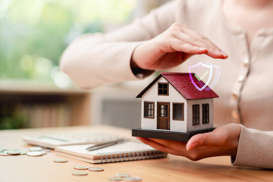 Home insurance, real estate protection and mortgage concept. Woman hands protecting miniature house model with safety shield icon, coins and calculator.