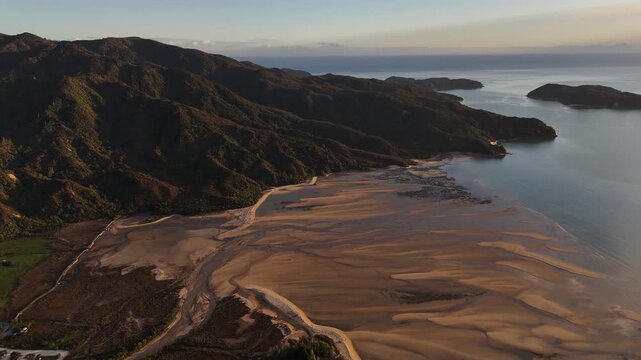 Coastal estuary with sweeping sand flats during low tide, surrounded by hills and vegetation at sunrise in Marahau, Abel Tasman National Park, New Zealand. Aerial drone