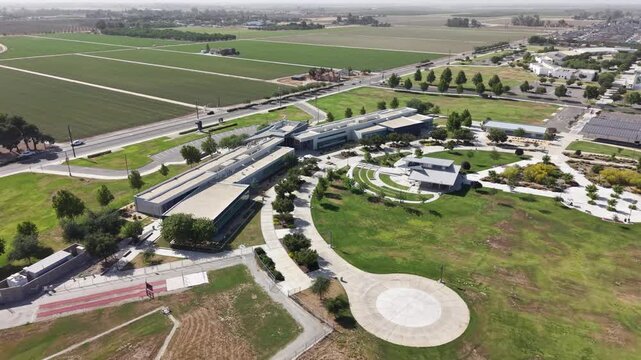Drone zoom out view of the College of the Sequoias campus in Visalia, California, showcasing modern architectural buildings surrounded by lush green lawns, manicured grounds, and agricultural fields
