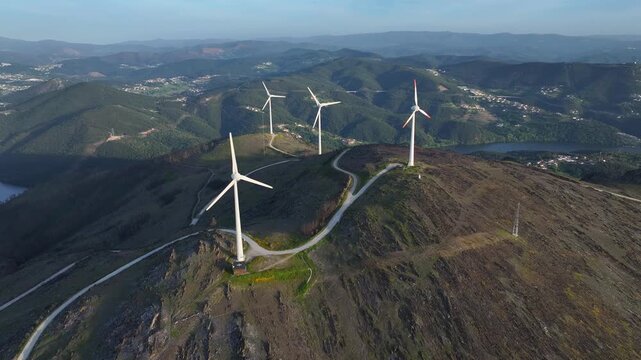 Wind turbines rotate on a hillside in a mountainous area. The farm generates clean energy. Surrounding landscapes include hills and a body of water.