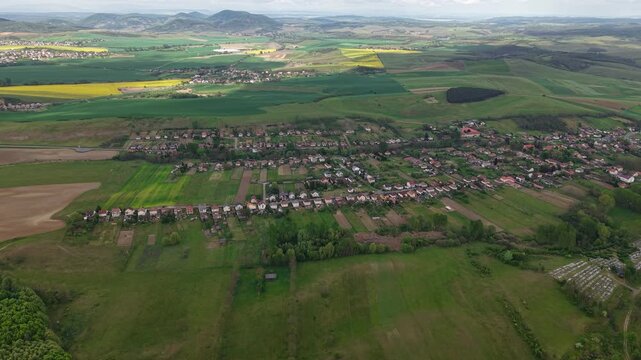 An expansive aerial perspective captures a tranquil village situated amidst a patchwork of vibrant green fields and agricultural land under a cloudy sky.