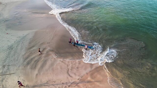 Fishermen unloading catch from fishing boat on sandy beach, fresh fish being handled on shore with coastal waves and local fishing activity in natural seaside environment overhead drone footage