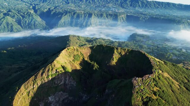 Mount Batur volcanic landscape with steep crater walls, drifting morning fog and glowing sunrise light across misty Bali highlands and forested mountain terrain push-in aerial drone shot