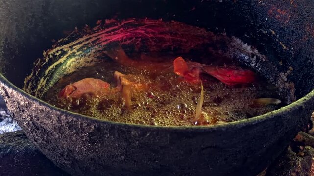Close-up of fresh fish frying in hot oil in large dark metal pot over open wood fire