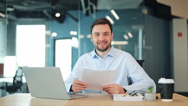 Smiling businessman or IT specialist analyzing papers or contracts at desk. Confident corporate worker, manager or freelance designer checking reports and looking at camera in coworking workspace.