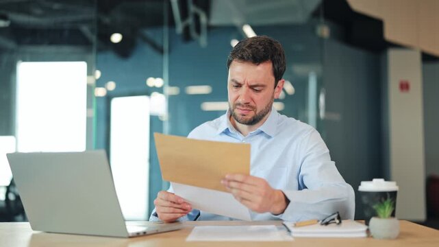 Focused businessman or IT specialist reading bad news or letter in craft envelope. Worried corporate worker, manager or freelance designer checking rejected contract or bill at modern desk.