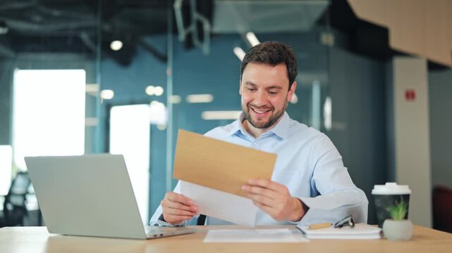Happy businessman or IT specialist reading good news or approved contract from craft envelope. Satisfied corporate worker, manager or freelance designer checking successful results at modern desk.