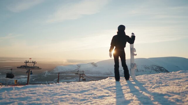 Slender woman snowboarder standing alone at top and admiring view of mountains. Rest in modern ski resort with infrastructure for skiing and snowboarding sports in winter, cable car for rising at top