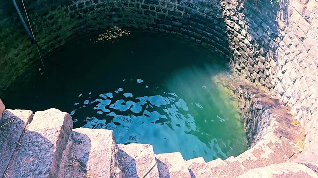 An ancient Indian dug well engineered with curved brick masonry. Stone steps descend down to the water table. Sunlight glints on the waving, rippled surface, revealing deep groundwater infrastructure.