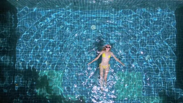 Top down drone view of woman floating in blue swimming pool during sunny tropical holiday and luxury resort relaxation.