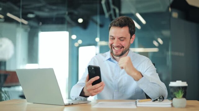 Close-up of happy businessman or IT specialist checking lottery win or financial growth on mobile phone. Satisfied corporate worker, manager or freelance designer celebrating success at office desk.