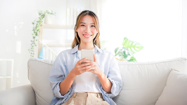 ソファでスマートフォンを持ちながらカメラ目線で微笑む若い女性
