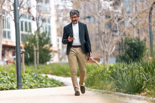 Happy mature businessman using his phone while walking through the park