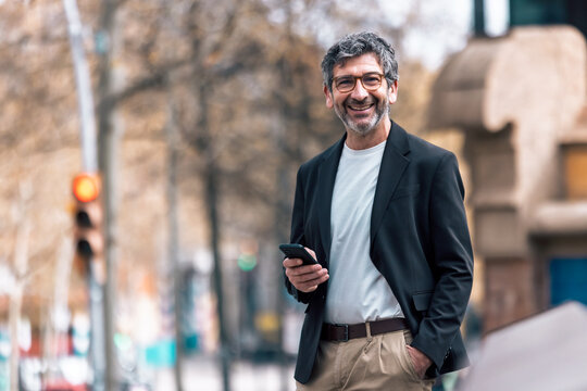 Happy mature elegant businessman using smartphone while walking through the city.