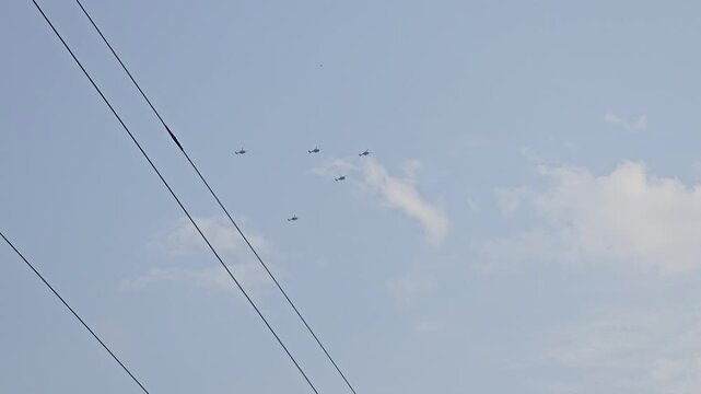 Five helicopters fly in formation above dark power lines, crossing into the open blue sky. The strategic aerial maneuver symbolizes national defense, vigilant security, and tactical operations.