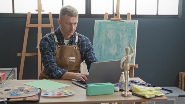 Man in apron dips brush with hand into water jar and paints on canvas beside laptop at table in studio; creative concentration.