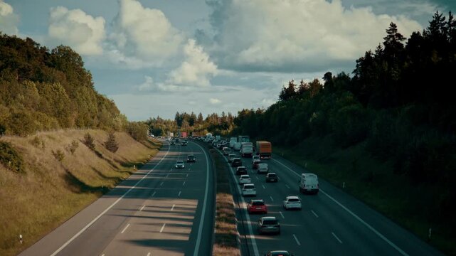 A high-angle view of a highway cutting through a green landscape where one side is congested with a long line of cars and trucks, while the other side flows freely under a cloudy sky.