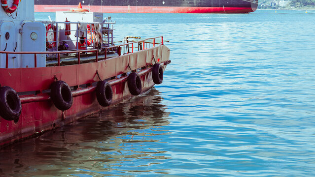 Red tugboat docked in calm blue water with a large cargo ship in the background.