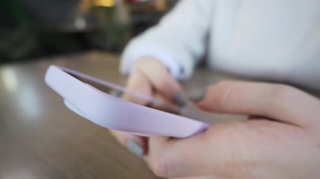 Female hands type on a smartphone with a purple case resting on a wooden cafe table. Concept of modern digital communication, social media networking, and online connectivity.