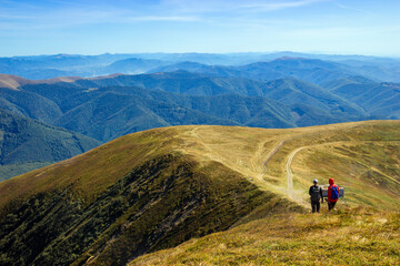 Obraz premium Two hikers walking along mountain ridge panoramic views rolling hills valleys under clear blue sky.
