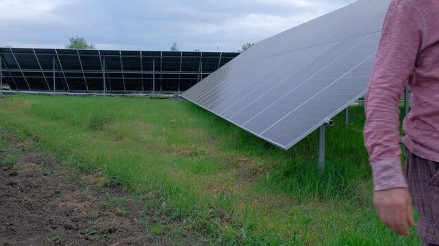Farmer checking solar panels performance while working in the garden A man with a hoe stands near home solar panels, calculating energy efficiency on a smartphone and taking notes in a notebook. 