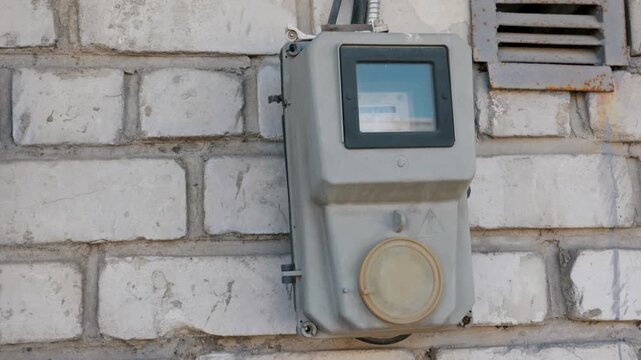 A close-up view of an old gray electrical power meter mounted securely on a textured white brick wall outside. Concept of energy consumption, utility costs, and electrical infrastructure.