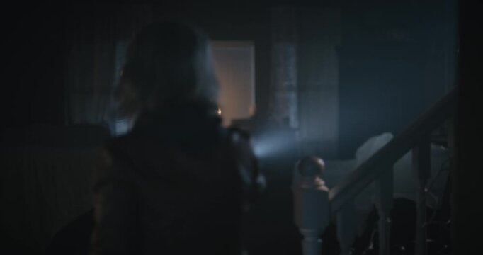 Dark silhouette of young woman searching dark haunted house with flashlight and startling herself - medium shot